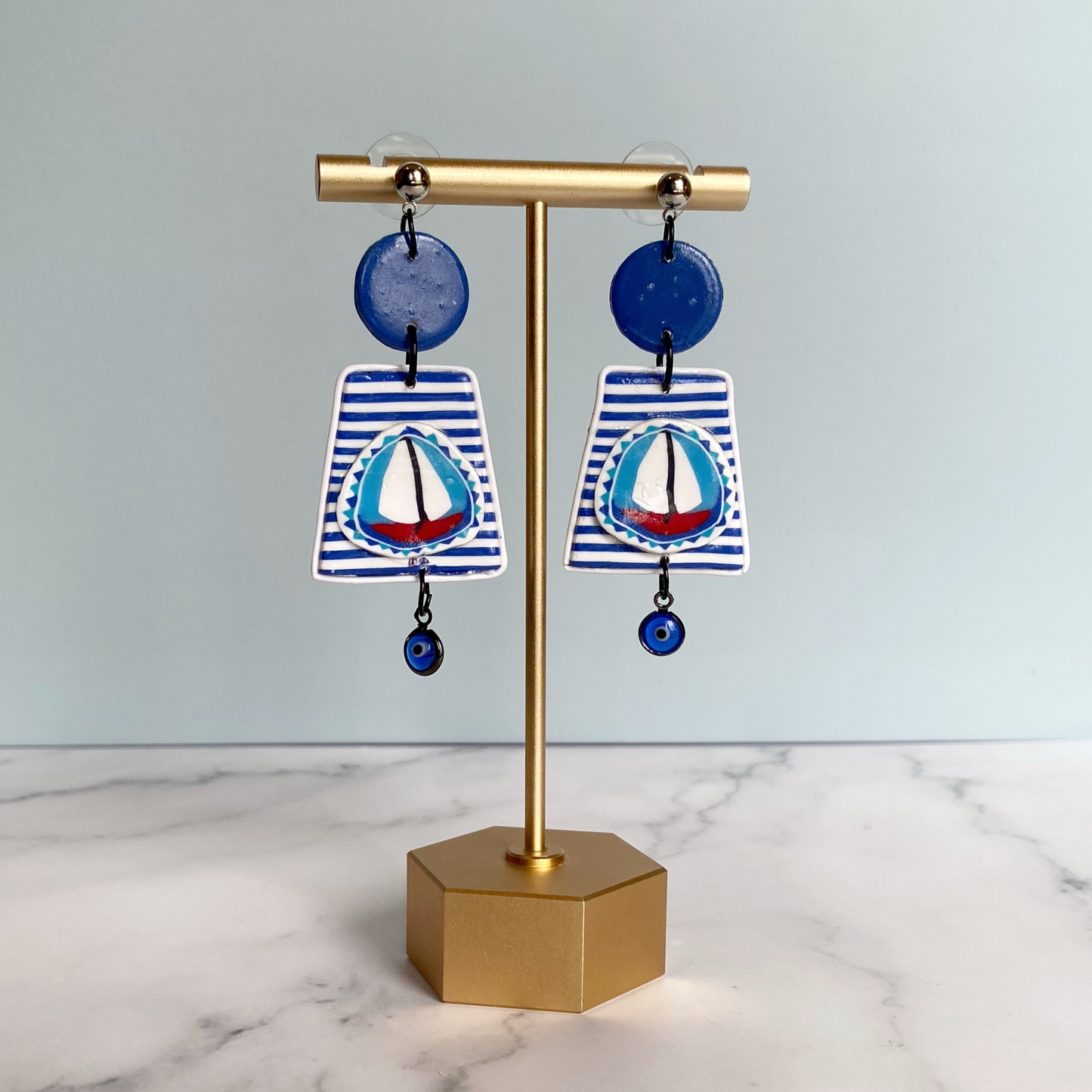 Blue and white striped handmade polymer clay sailboat earrings on a brass earring stand.