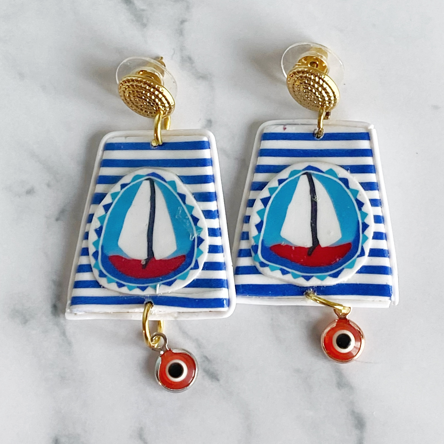 Handmade Polymer Clay Earrings with a blue and white striped background and a red sailboat with white sails on a marble background.