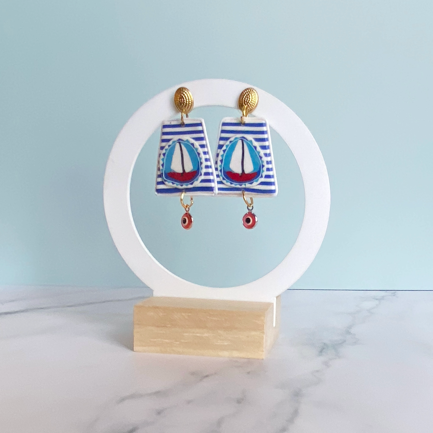 Handmade Polymer Clay Earrings with a blue and white striped background and a red sailboat with white sails on a white circular stand.