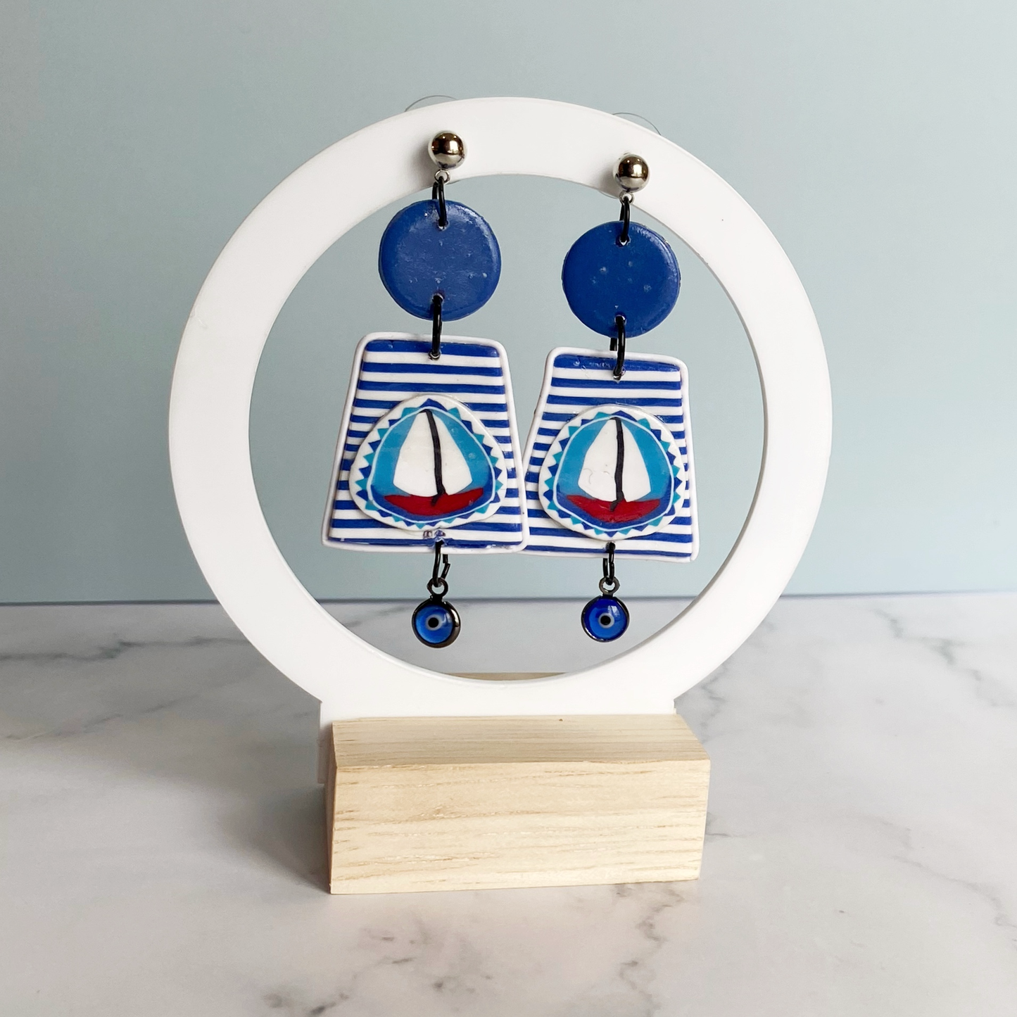 Blue and white striped handmade polymer clay sailboat earrings on a chite circle stand.
