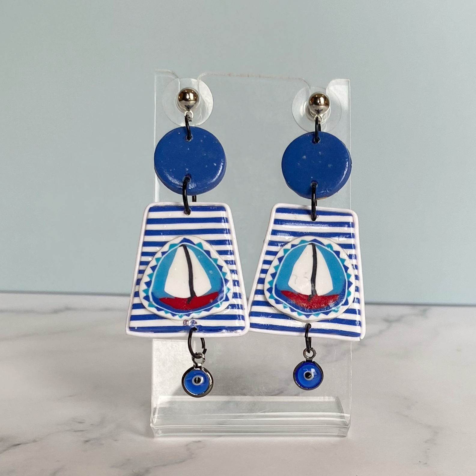 Blue and white striped handmade polymer clay sailboat earrings on a clear acrylic stand.