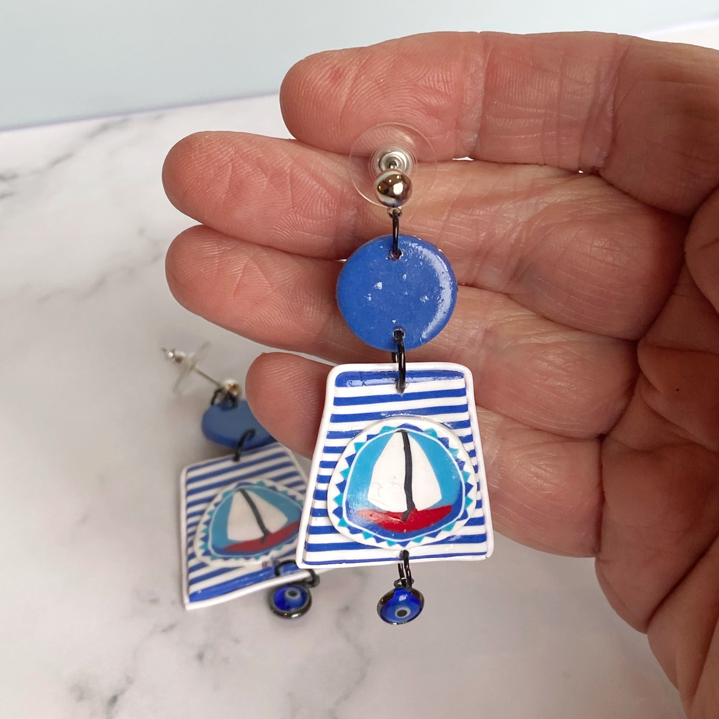 Blue and white striped handmade polymer clay sailboat earrings handheld for size reference.