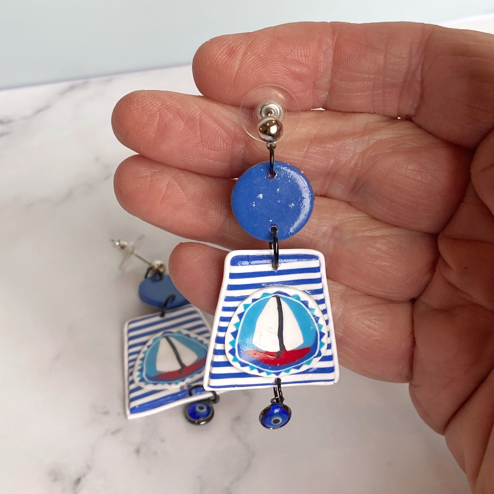 Blue and white striped handmade polymer clay sailboat earrings handheld for size reference.